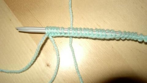 Wenn du Strickmaschen aufnehmen möchtest, hilft dir diese Anleitung, den ersten Schritt richtig zu machen. Das Bild zeigt eine klare Übersicht über den Anfang deines Strickprojekts mit einer neuen Masche. Eine Stricknadel mit grüner Wolle ist auf einem Holzuntergrund zu sehen, während Maschen aufgenommen werden.