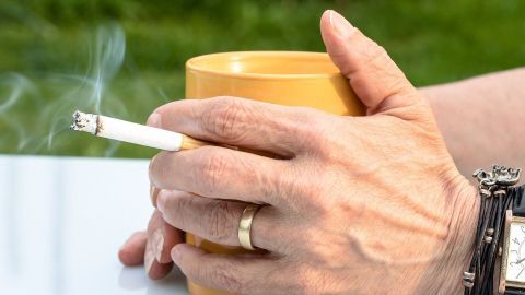 Eine Hand hält eine Zigarette über einem gelben Becher, während im Hintergrund grünes Gras sichtbar ist. Nikotinflecken und Hautverfärbung sind Thema des Bildes.