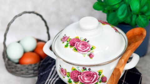 Ein dekorativer Topf mit Rosenmuster steht auf einem Tuch, während frische Eier in einem Korb daneben liegen.
