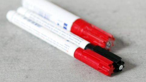 Mehrere Edding-Stifte liegen auf einer grauen Oberfläche. Zwei Kappen sind rot, eine ist schwarz, alle Stifte sind weiß.