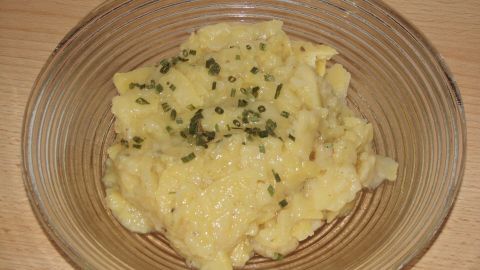 Ein Kartoffelsalat liegt in einer Glasschüssel, garnisiert mit Kräutern, auf einem Holztisch. Schwäbischer Kartoffelsalat ist bereit zum Servieren.
