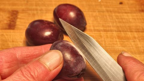 Zwetschgen entkernt und braune Hände bekommen? Die Flecken lassen sich gut mit Essigwasser entfernen. Eine Hand hält eine Pflaume, während ein Messer die Schale abzieht; die Holzoberfläche bildet den Hintergrund.