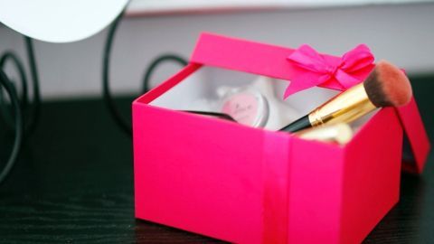 Schminkschachtel mit Pinsel in einer pinken Geschenkbox steht geöffnet auf einem Tisch, geschmückt mit einer Schleife.