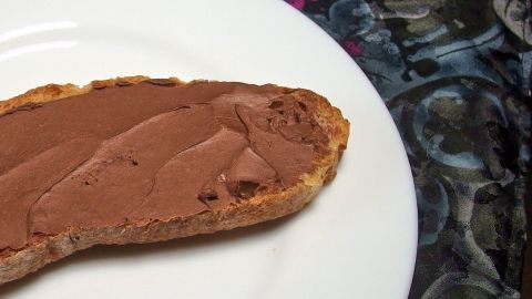 Ein geröstetes Brotstück mit samtiger Haselnusscreme liegt auf einem weißen Teller vor einem bunten, gemusterten Hintergrund.