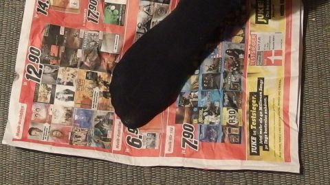 Ein schwarzer Fußsocken liegt auf einer ausgebreiteten Zeitungsseite, die auf einem Teppichboden platziert ist.