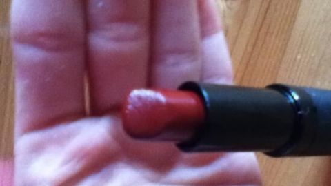 Ein geöffneter, roter Lippenstift wird von einer Hand gehalten, im Hintergrund ist ein Holztisch zu sehen.