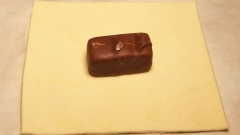 Superleckerer Blätterteig mit Celebrations- (Mars, Snickers, Milky Way, etc.) - ganz schnell und einfach zubereitet. Ein Stück Blätterteig liegt auf einer Arbeitsfläche, während ein Schokoladenstück darauf platziert ist, bereit zum Falten.