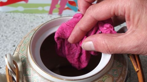Eine Hand hält ein pinkes Tuch, das in eine Teekanne mit Verfärbungen eingetaucht wird, um Kaffee- und Teeränder zu entfernen.