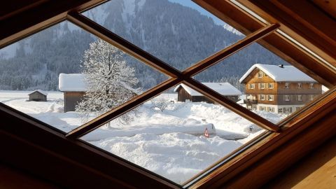 Ein Fenster zeigt eine verschneite Landschaft mit einem Haus und Bäumen. Umgezogen, jedoch ohne Sichtschutz am Fenster.