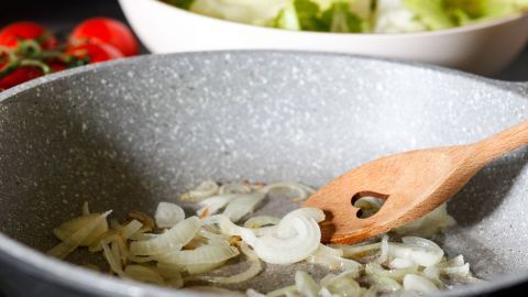 In einer Pfanne braten Zwiebeln an, während im Hintergrund frischer Salat und Tomaten auf einem Tisch stehen.