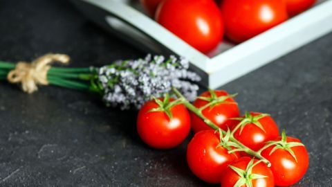 Saftige, gekochte Tomaten liegen neben einem Bündel frischer Lavendel auf einer dunklen Oberfläche, umgeben von weiteren Tomaten.