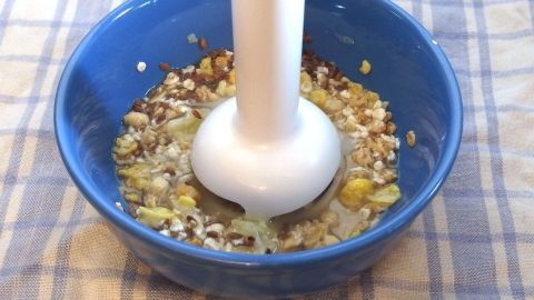 Ein blauer Schüssel enthält Müsli, während ein Stabmixer es zu einer trinkbaren Konsistenz verarbeitet.