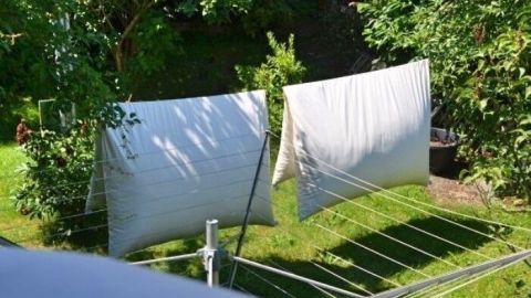 Weiße Bettwäsche hängt frisch gewaschen auf einer Wäscheleine im Garten, um in der Sonne zu trocknen.