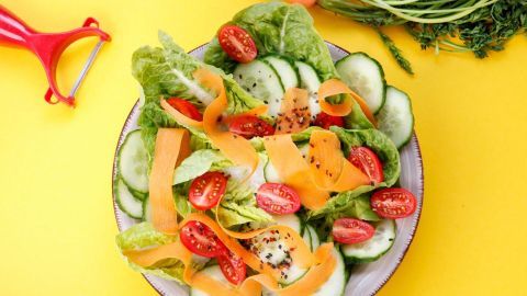 Frischer Salat mit Möhrenbandnudeln und Tomaten, angerichtet in einer Schüssel auf gelbem Hintergrund.