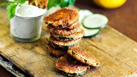 Panierte Zucchini liegen gestapelt auf einem Holzbrett, daneben Minz-Dip in einem kleinen Glas und frische Zitronen.
