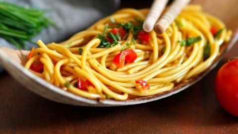 Spaghetti auf einem Holzlöffel, garniert mit grünen Kräutern und roten Paprikawürfeln, daneben eine frische Tomate.