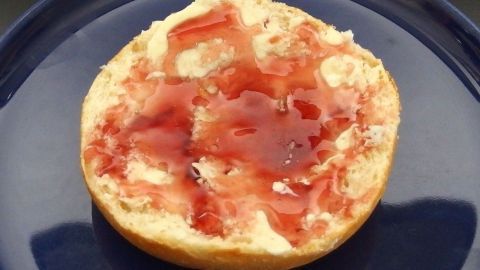 Ein Brötchen mit rotem Holunder Gelee liegt auf einem blauen Teller und sorgt für ein einladendes Frühstücksgefühl.