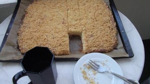 Ein Butter-Streusel-Kuchen mit Frischkäse liegt auf einem Tablett, umgeben von einer Tasse und einem leeren Teller.