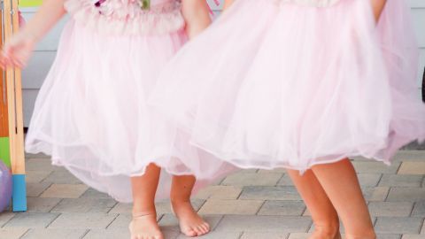 Zwei Mädchen in rosa Tüllkleidern tanzen barfuß auf einem gepflasterten Boden in einer fröhlichen Umgebung.