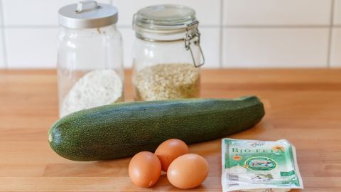 Heute stelle ich euch mal das Zucchini-Burger-Rezept vor, für etwa 700 g Zucchini. Auf einem Holztisch liegen eine Zucchini, drei Eier, Haferflocken und eine Packung Käse, bereit für einen vegetarischen Zucchini-Burger.