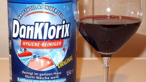 Ein Behälter mit DanKlorix steht neben einem Glas Rotwein, das auf einem Tisch platziert ist. Der Fokus liegt auf der Reinigungsmittelpackung.