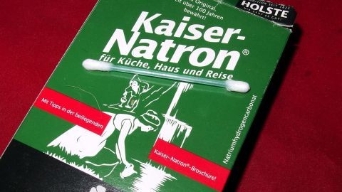 Eine grüne Verpackung von Kaiser-Natron liegt auf einem roten Untergrund, bereit zur Verwendung gegen schmerzhafte Aphten.