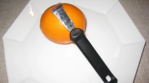 Eine Zestenreißer liegt auf einem weißen Teller, während sie die Schale einer orangefarbenen Orange abzieht.