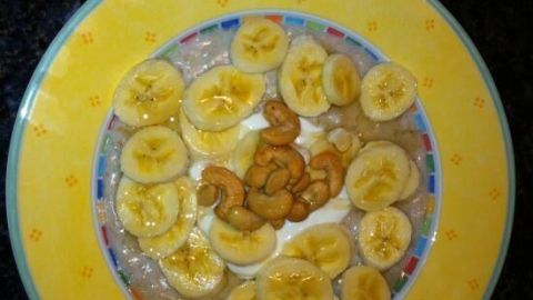 Ein kleiner Teller mit Porridge ist mit geschnittenen Bananen und gesalzenen Cashewkernen dekoriert, auf dunklem Hintergrund.