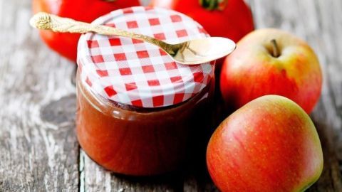 Ein Glas Apfel-Tomaten-Chutney, dekoriert mit einem karierten Deckel, steht auf einem rustikalen Holztisch neben frischen Äpfeln und Tomaten.