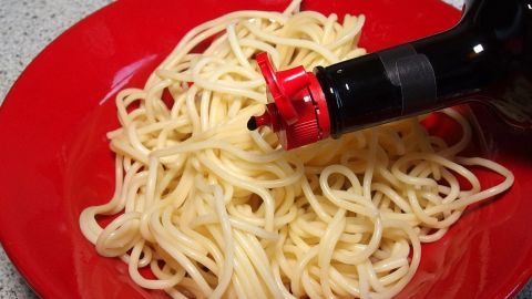 Ein roter Teller mit Spaghetti wird mit Maggi-Würze übergossen, während die Nudeln auf einem Tisch serviert werden.