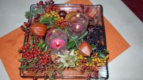 Eine dekorative Glasplatte mit Kerzen und herbstlichen Früchten und Beeren liegt auf einem orangefarbenen Untersetzer.