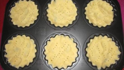 Sechs Mürbeteigförmchen, bereit zum Backen, stehen in einer dunklen Muffinform auf einem pinken Untergrund.