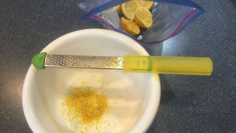 Eine Zesterreibe liegt auf einer Schüssel, während Zitronenschale darin gerieben wird, auf einer Tischoberfläche mit einem Zipbeutel daneben.
