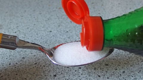 Eine Esslöffel mit Zucker wird aus einer Flasche mit rotem Deckel gefüllt, auf einer grauen Küchenoberfläche.