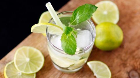 Ein Glas Virgin Caipirinha mit Limettenscheiben und Minze auf einem Holzbrett, umgeben von weiteren Limetten.
