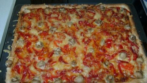 Pizza mit Pilzen und Paprika liegt gebacken auf einem schwarzen Backblech in einer Küche.