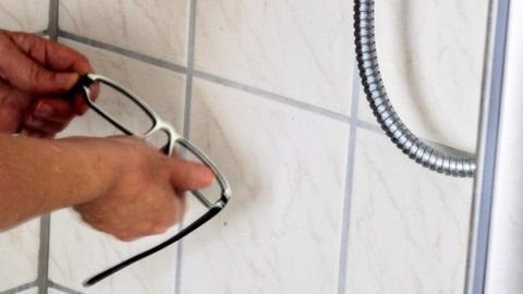 Eine Person hält eine Brille und reinigt sie unter einem Duschkopf mit Haarshampoo, um sie zu säubern.