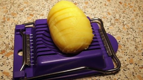 Eine geschälte Kartoffel liegt auf einem lila Eierschneider, bereit zum Schneiden für Bratkartoffeln auf einer Arbeitsfläche.