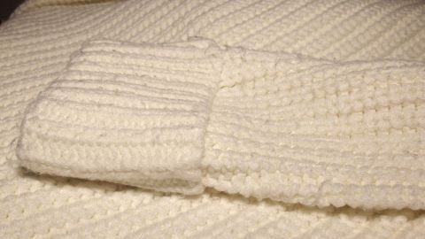 Kratzende Pullover sind sehr unangenehm zu tragen. Lies jetzt, was man dagegen machen kann. Ein kratzender Pullover liegt auf einem Tisch, dessen Struktur grob und sehr dick ist, in einer neutralen Umgebung.