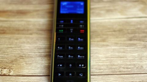 Ein drahtloses Telefon mit beleuchtetem Display liegt auf hölzernem Untergrund. Das Display zeigt unleserlichen Text.