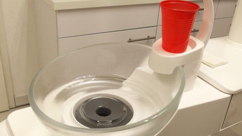 Eine transparente Schüssel mit einer zentralen Öffnung steht in einem modernen Bad, während ein roter Becher darauf platziert ist.