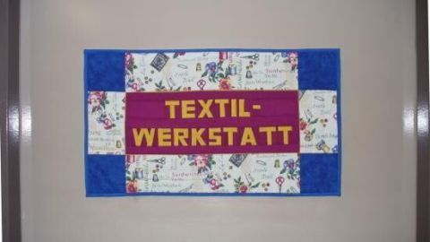 Ein buntes Schild mit "TEXTILWERKSTATT" hängt an einer Wand. Es besteht aus Stoff mit floralen und geometrischen Mustern.