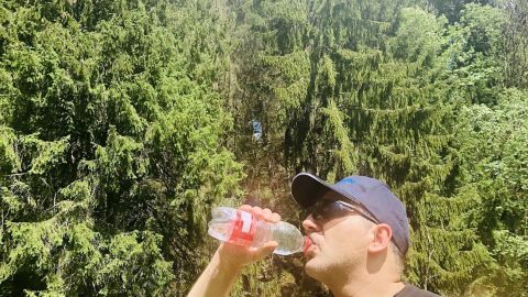 Ein Mann trinkt aus einer Flasche, während er in einem Wald mit hohen Bäumen steht und die Sonne scheint.