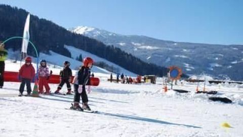 Skifahrende Kinder üben auf der verschneiten Piste, umgeben von Bergen und anderen Familien im Alpenurlaub.
