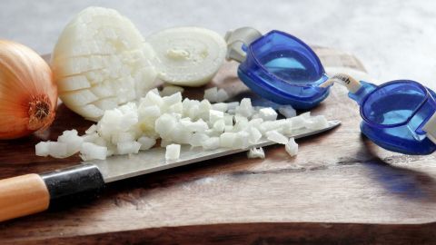 Eine halbierte Zwiebel liegt neben gewürfelten Zwiebelstücken auf einem Holzbrett, während ein Messer und Schutzbrille bereitstehen.