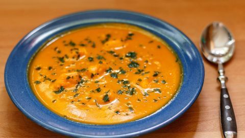 Eine orange Kürbiscremesuppe steht in einer blauen Schale, dekoriert mit Kräutern, neben einem Löffel auf einem Holztisch.