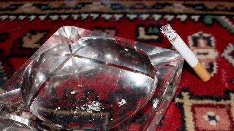 Ein Aschenbecher steht auf einem bunten Teppich, während eine brennende Zigarette daneben liegt, die Brandflecken hinterlassen könnte.