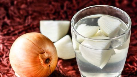 Eine Zwiebel liegt neben einem Glas mit Zwiebelwasser, bereit zum Gurgeln gegen Halsschmerzen, auf einem roten Hintergrund.