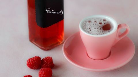 Eine rosa Tasse steht auf einem Teller, während Himbeeressig in einer Flasche daneben platziert ist; frische Himbeeren liegen daneben.