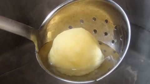 Ein Kartoffelknödel liegt in einem Sieblöffel, während er in einem Topf mit kochendem Wasser gegart wird.